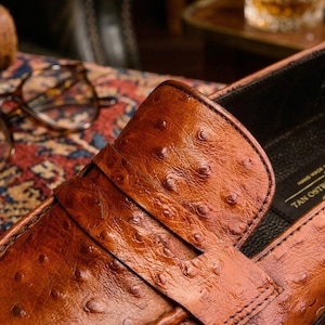 K&ouml;nnte beinhalten: Nahaufnahme eines braunen Lederloafers mit strukturierter Oberfl&auml;che. Der Schuh hat einen Riemen &uuml;ber der Oberseite und ist mit "TAN OSTRICH" und "HAND MADE IN ITALY" beschriftet. Der Schuh ist vor einem unscharfen Hintergrund platziert.