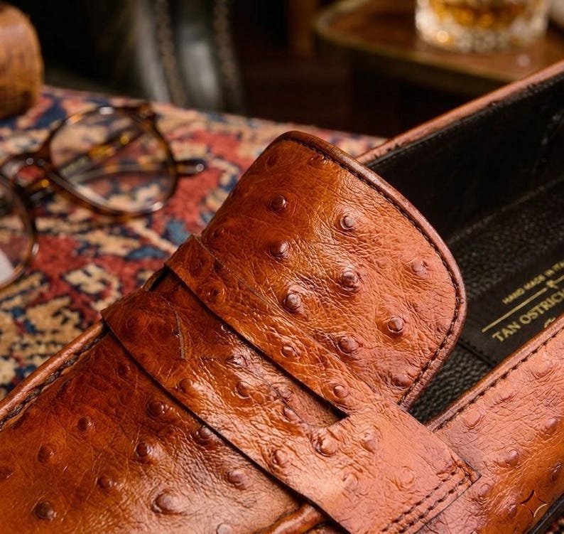 K&ouml;nnte beinhalten: Nahaufnahme eines braunen Lederloafers mit strukturierter Oberfl&auml;che. Der Schuh hat einen Riemen &uuml;ber der Oberseite und ist mit "TAN OSTRICH" und "HAND MADE IN ITALY" beschriftet. Der Schuh ist vor einem unscharfen Hintergrund platziert.