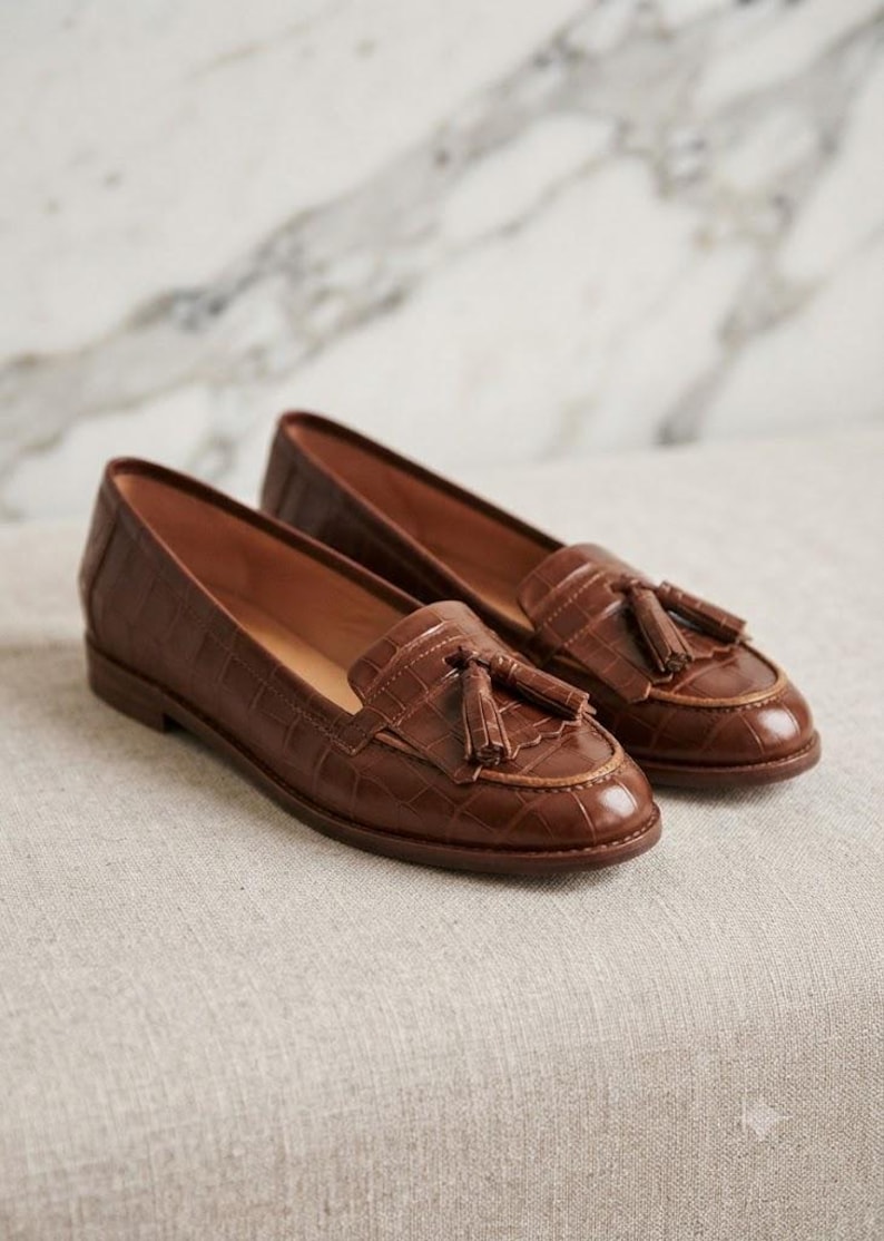 May include: A pair of brown leather loafers featuring a crocodile-embossed texture, a rounded toe, and a decorative tassel detail on the vamp. The shoes have a low heel and a smooth interior lining, showcasing a classic slip-on design.