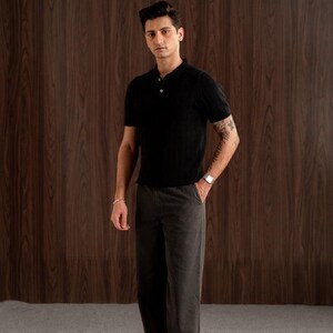 May include: A person wearing a black ribbed polo shirt with three buttons and dark grey trousers. The outfit is completed with a silver watch and bracelet. The background is a wooden wall and a light-coloured rug.