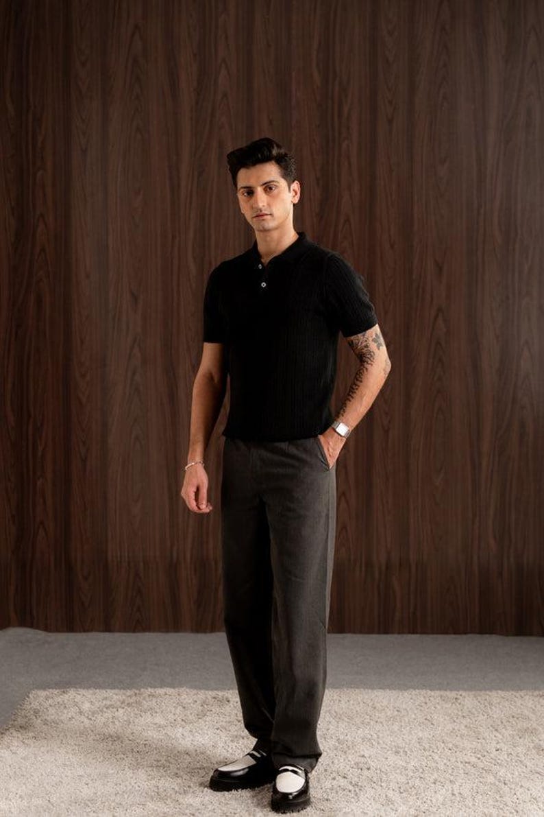 May include: A person wearing a black ribbed polo shirt with three buttons and dark grey trousers. The outfit is completed with a silver watch and bracelet. The background is a wooden wall and a light-coloured rug.