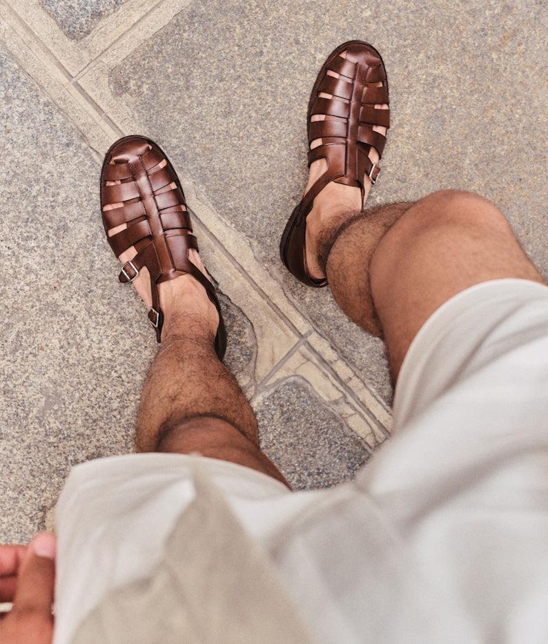 May include: A pair of brown leather sandals with a woven design. The sandals have a closed-toe design with a buckle closure. The sandals are worn on a person's feet, with the legs and white shorts visible.