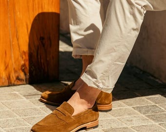 May include: Brown suede penny loafers with a classic design. The shoes have a rounded toe, a leather sole, and a strap across the vamp. The loafers are worn with beige pants. The shoes are a stylish choice for casual or semi-formal occasions.