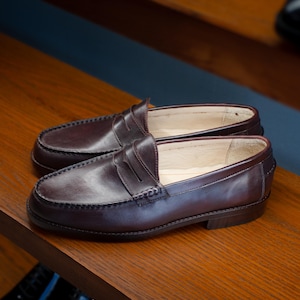 May include: A pair of dark brown leather penny loafers on a wooden shelf. The shoes have a classic design with a rounded toe, a strap across the vamp, and a low heel. The leather appears smooth and polished.