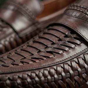May include: Close-up of brown leather loafers with woven detailing. The shoes have a classic design with a rounded toe and a slip-on style. The leather appears to be high-quality, with a rich, dark brown color and visible texture.