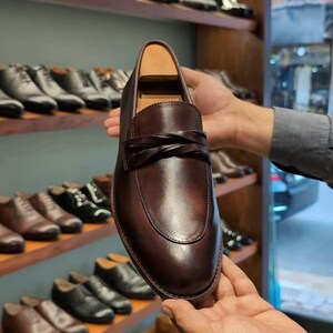 Puede incluir: Primer plano de un mocasín de cuero marrón con un detalle de correa trenzada. El zapato se sostiene frente a una exhibición de otros zapatos de cuero en estantes de madera. El mocasín tiene un acabado pulido.