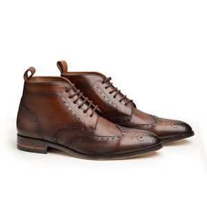 Könnte beinhalten: Ein Paar braune Leder-Ankle Boots mit Brogue-Details. Die Stiefel haben ein klassisches Design mit Schnürverschluss und kleinem Absatz. Das Leder hat eine polierte Oberfläche, vor weißem Hintergrund.