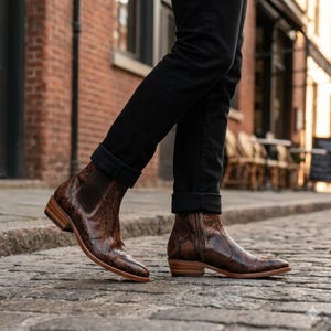May include: Brown leather ankle boots with a snakeskin pattern. The boots have a pointed toe, a side zipper, and a wooden heel. The boots are worn with black jeans. The boots are a stylish footwear choice.