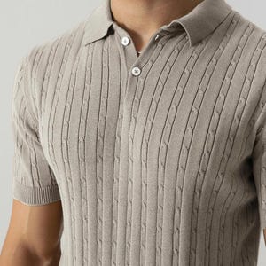 May include: A light beige polo shirt featuring a cable knit pattern. The short-sleeved top has a collared neckline and three buttons. The material looks soft and breathable.