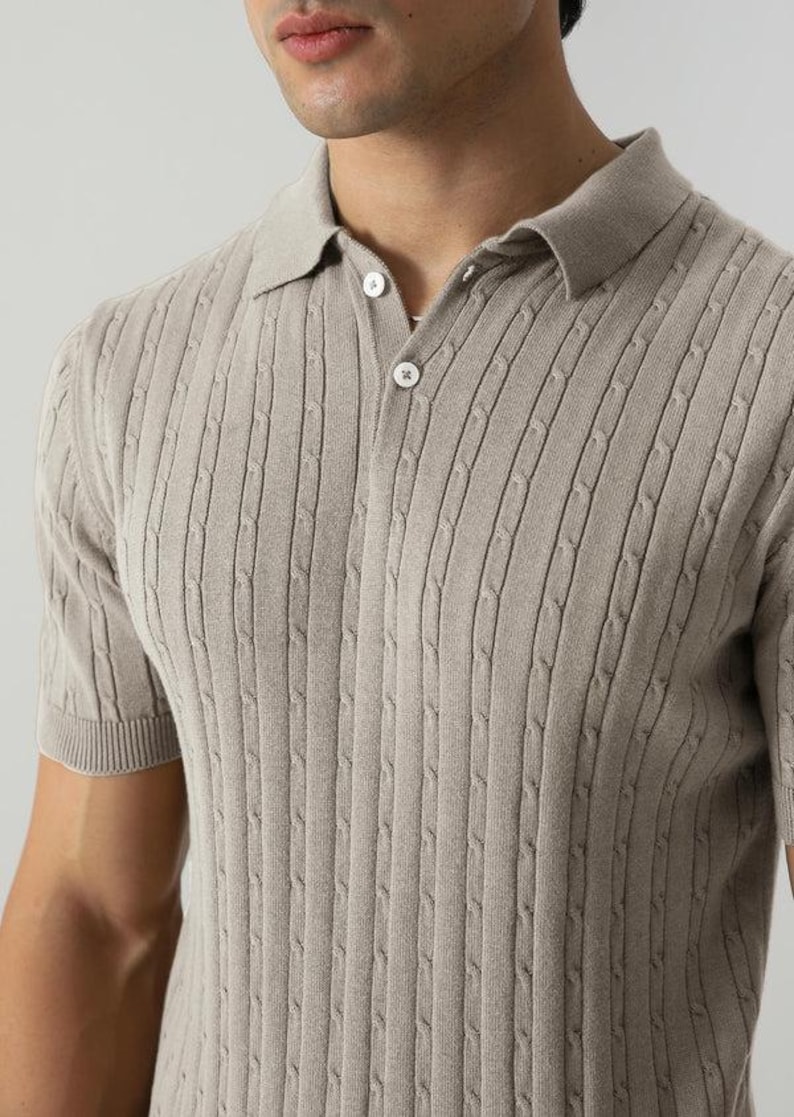 May include: A light beige polo shirt featuring a cable knit pattern. The short-sleeved top has a collared neckline and three buttons. The material looks soft and breathable.