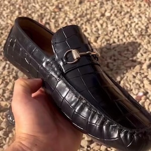 May include: Black leather loafer with a crocodile texture. The shoe features a silver-colored metal buckle detail on the vamp. The loafer is held in a hand, showcasing its glossy finish and design.