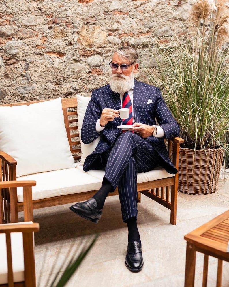 Pu&ograve; includere: Una persona in un abito gessato blu navy con cravatta a righe rosse e blu, scarpe nere e occhiali da sole &egrave; seduta su una panchina di legno, tenendo in mano una tazza di caff&egrave;. Lo sfondo presenta un muro di mattoni e erba alta.