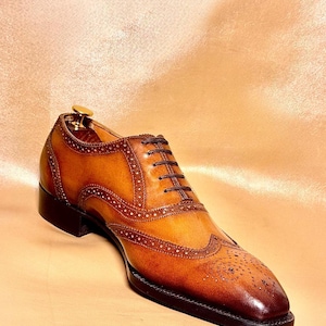 Puede incluir: Un zapato Oxford de cuero marr&oacute;n con detalles brogue. El zapato presenta una suela y un tac&oacute;n marr&oacute;n oscuro, con una parte superior marr&oacute;n m&aacute;s clara. Los cordones son de color marr&oacute;n oscuro. El zapato se muestra sobre una superficie marr&oacute;n claro.