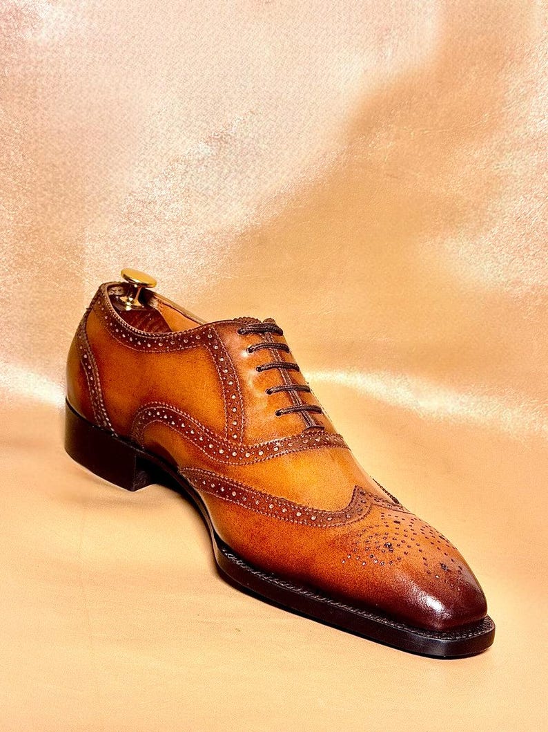 Puede incluir: Un zapato Oxford de cuero marr&oacute;n con detalles brogue. El zapato presenta una suela y un tac&oacute;n marr&oacute;n oscuro, con una parte superior marr&oacute;n m&aacute;s clara. Los cordones son de color marr&oacute;n oscuro. El zapato se muestra sobre una superficie marr&oacute;n claro.