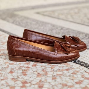 May include: A pair of brown leather loafers featuring a crocodile-embossed texture, a rounded toe, and a decorative tassel detail on the vamp. The shoes have a low, flat heel and a smooth interior lining, showcasing a classic slip-on construction.