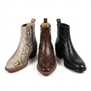 May include: Three ankle boots in a row, each with a snakeskin pattern. The boots are in shades of light grey, brown, and black. Each boot has a zipper on the side and a small heel. The boots are displayed on a white background.