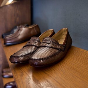 May include: A pair of brown loafers with a textured, woven-like pattern on the upper surface. The shoes feature a classic penny loafer strap across the vamp, a rounded toe, and a smooth interior lining, displayed on a wooden shelf.