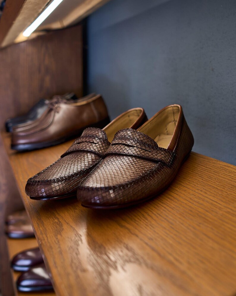 May include: A pair of brown loafers with a textured, woven-like pattern on the upper surface. The shoes feature a classic penny loafer strap across the vamp, a rounded toe, and a smooth interior lining, displayed on a wooden shelf.