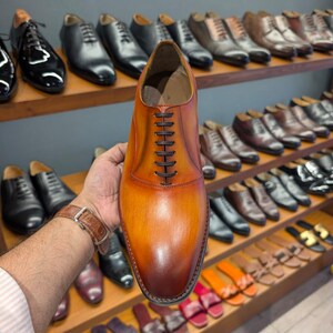 May include: A brown leather dress shoe with dark brown laces is held up in front of a display of other shoes. The shoe has a gradient color, transitioning from light brown to a darker shade. Other shoes are visible on wooden shelves.