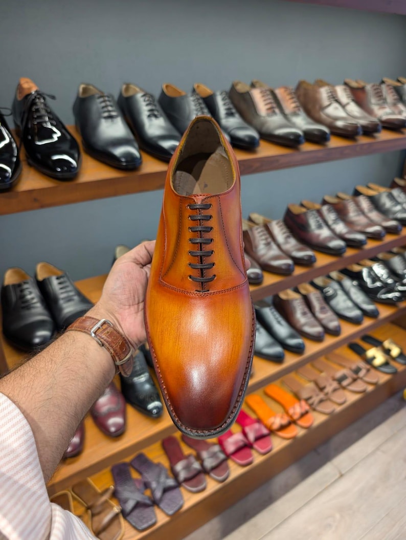 May include: A brown leather dress shoe with dark brown laces is held up in front of a display of other shoes. The shoe has a gradient color, transitioning from light brown to a darker shade. Other shoes are visible on wooden shelves.