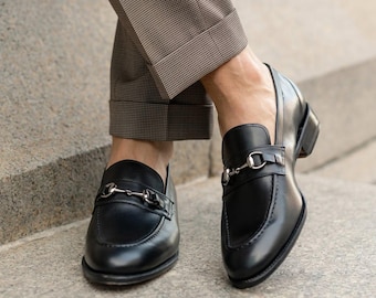 May include: Black leather loafers with silver-tone metal horsebit detail. The shoes have a low heel and a classic design. The image shows the shoes on a person's feet, paired with brown patterned trousers.