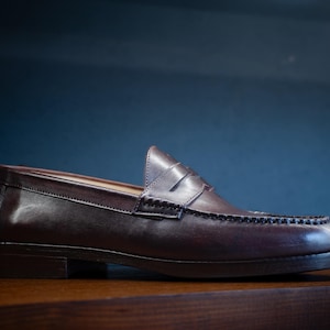 May include: A brown leather loafer shoe with a penny strap and decorative stitching. The shoe is made of polished leather and sits on a wooden surface. The background is a dark blue.