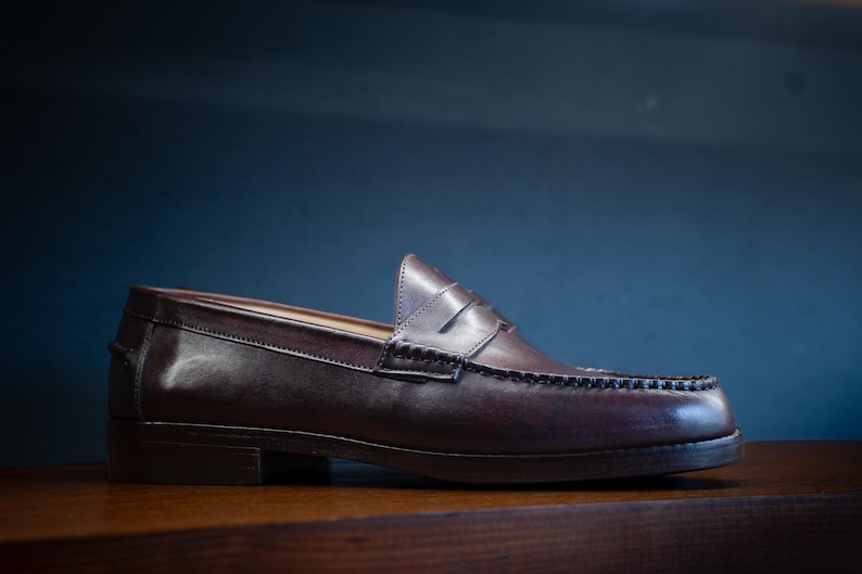 May include: A brown leather loafer shoe with a penny strap and decorative stitching. The shoe is made of polished leather and sits on a wooden surface. The background is a dark blue.