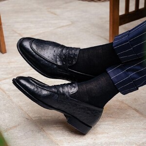 Pu&ograve; includere: Mocassini in pelle nera con finitura strutturata. Le scarpe hanno un design classico con cinturino penny sulla tomaia. La persona indossa calzini neri e pantaloni a righe blu. Calzature eleganti.