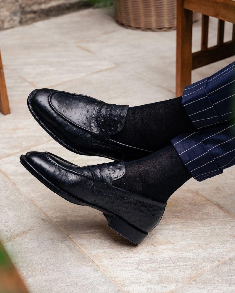 Pu&ograve; includere: Mocassini in pelle nera con finitura strutturata. Le scarpe hanno un design classico con cinturino penny sulla tomaia. La persona indossa calzini neri e pantaloni a righe blu. Calzature eleganti.