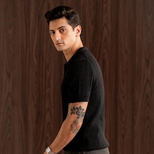 May include: A black short-sleeved polo shirt with a textured knit pattern. The shirt is paired with grey trousers. The person is wearing a silver watch and has a tattoo on their arm. The background is a dark wood panel.