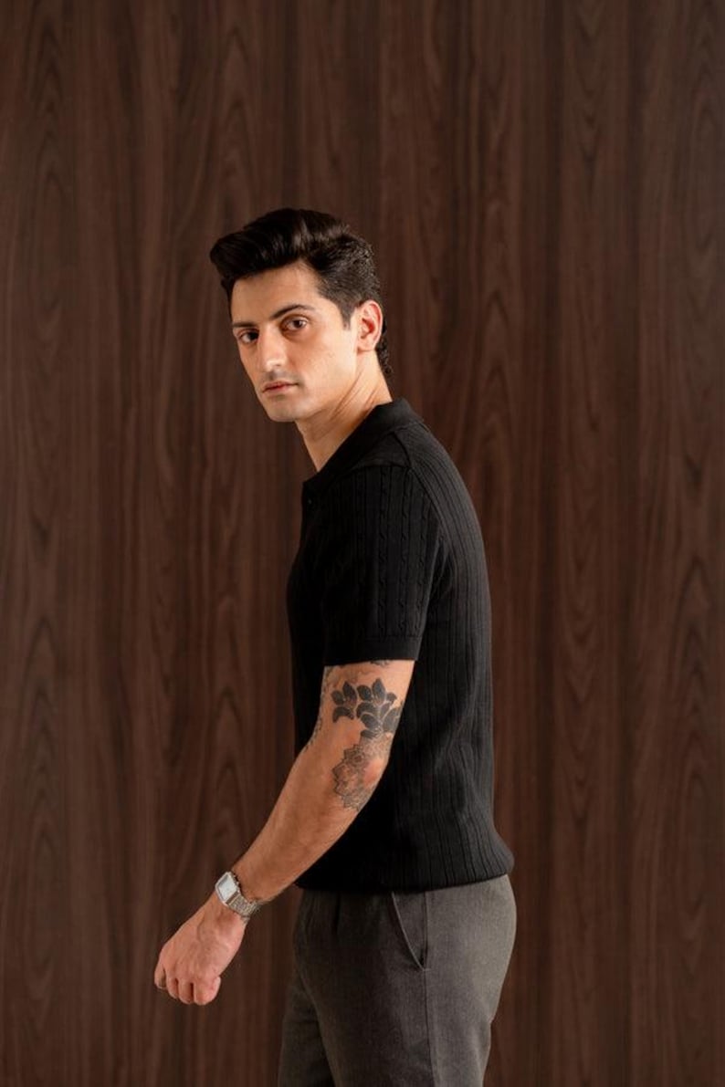 May include: A black short-sleeved polo shirt with a textured knit pattern. The shirt is paired with grey trousers. The person is wearing a silver watch and has a tattoo on their arm. The background is a dark wood panel.