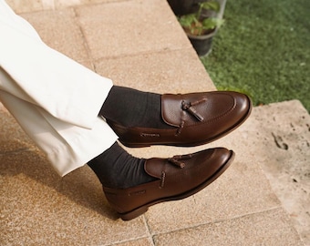 May include: A pair of brown leather loafers with tassel details. The shoes are a rich, dark brown color and appear to be made of textured leather. The loafers are worn with black socks and white pants.