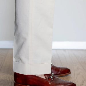 May include: A pair of polished, dark brown leather loafers with silver-colored metal accents. The shoes are paired with light beige trousers. The image shows the lower legs and feet of a person standing on a wooden floor.