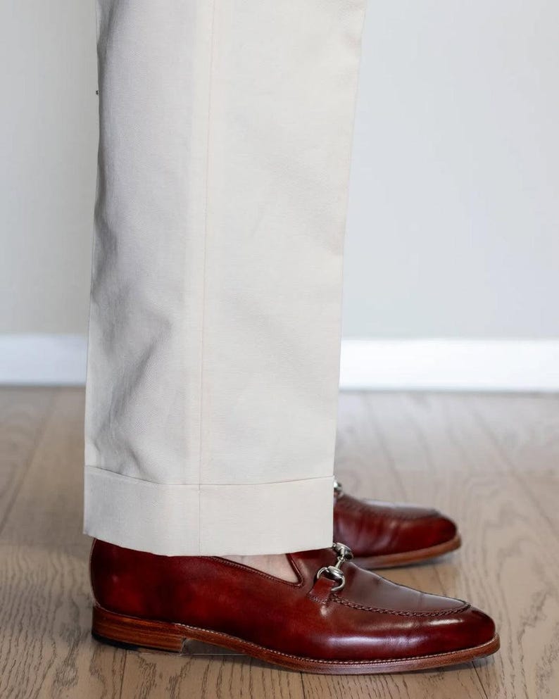 May include: A pair of polished, dark brown leather loafers with silver-colored metal accents. The shoes are paired with light beige trousers. The image shows the lower legs and feet of a person standing on a wooden floor.