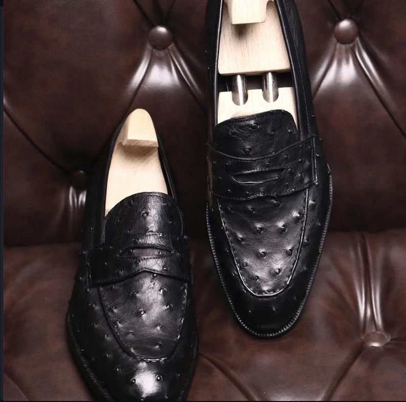 Pu&ograve; includere: Un paio di mocassini neri in pelle con una superficie strutturata, caratterizzati da un classico design penny loafer. Le scarpe hanno le forme in legno inserite. I mocassini sono esposti su una superficie in pelle marrone.