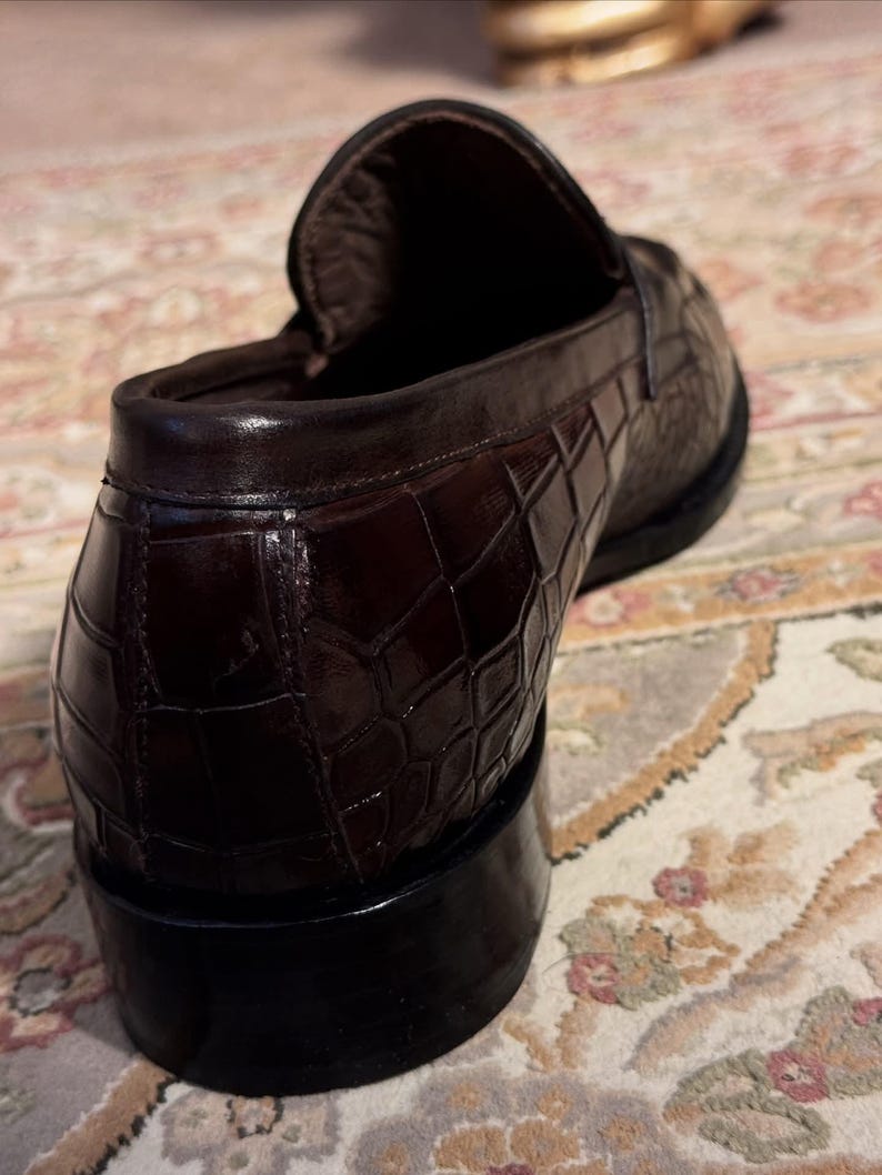 Rear view of a men's burgundy crocodile embossed leather loafer showing the sturdy stacked heel, reinforced heel counter, and detailed scale texture against a patterned rug.