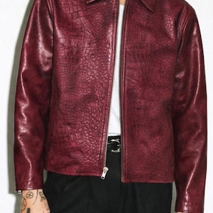 May include: A burgundy faux leather jacket with a textured, crocodile-like pattern. The jacket has a collared neckline, a silver zipper, and long sleeves. The model is wearing a white t-shirt underneath and black pants.