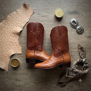 Puede incluir: Un par de botas vaqueras de cuero marrón con un empeine de piel de avestruz texturizada y una caña de cuero liso con costuras decorativas. Las botas se muestran junto a un trozo de piel de avestruz, una lata redonda, una hebilla y un pañuelo.