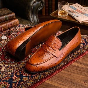 K&ouml;nnte beinhalten: Braune Leder-Loafer mit strukturierter Oberfl&auml;che. Die Schuhe haben ein klassisches Design zum Hineinschl&uuml;pfen und einen niedrigen Absatz. Sie werden auf einem gemusterten Teppich pr&auml;sentiert. Die Schuhe sind ca. 28 cm lang.