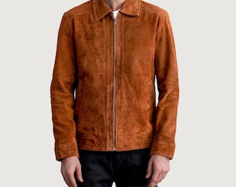 May include: A brown suede jacket with a collared neckline and a full-length zipper. The jacket has long sleeves and a classic design. The model is wearing a white t-shirt underneath and dark blue jeans.