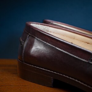 May include: Close-up of a pair of brown leather loafers. The shoes have a classic design with a smooth, polished finish. The image highlights the stitching and the quality of the leather. The loafers are on a wooden surface.