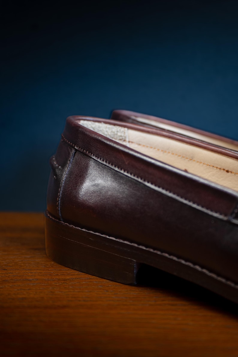 May include: Close-up of a pair of brown leather loafers. The shoes have a classic design with a smooth, polished finish. The image highlights the stitching and the quality of the leather. The loafers are on a wooden surface.