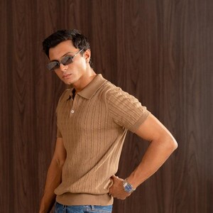 May include: A tan short-sleeved polo shirt with a textured cable knit design. The shirt has a collared neckline and a button closure. The model is wearing blue jeans and sunglasses. A watch is on the model's wrist.