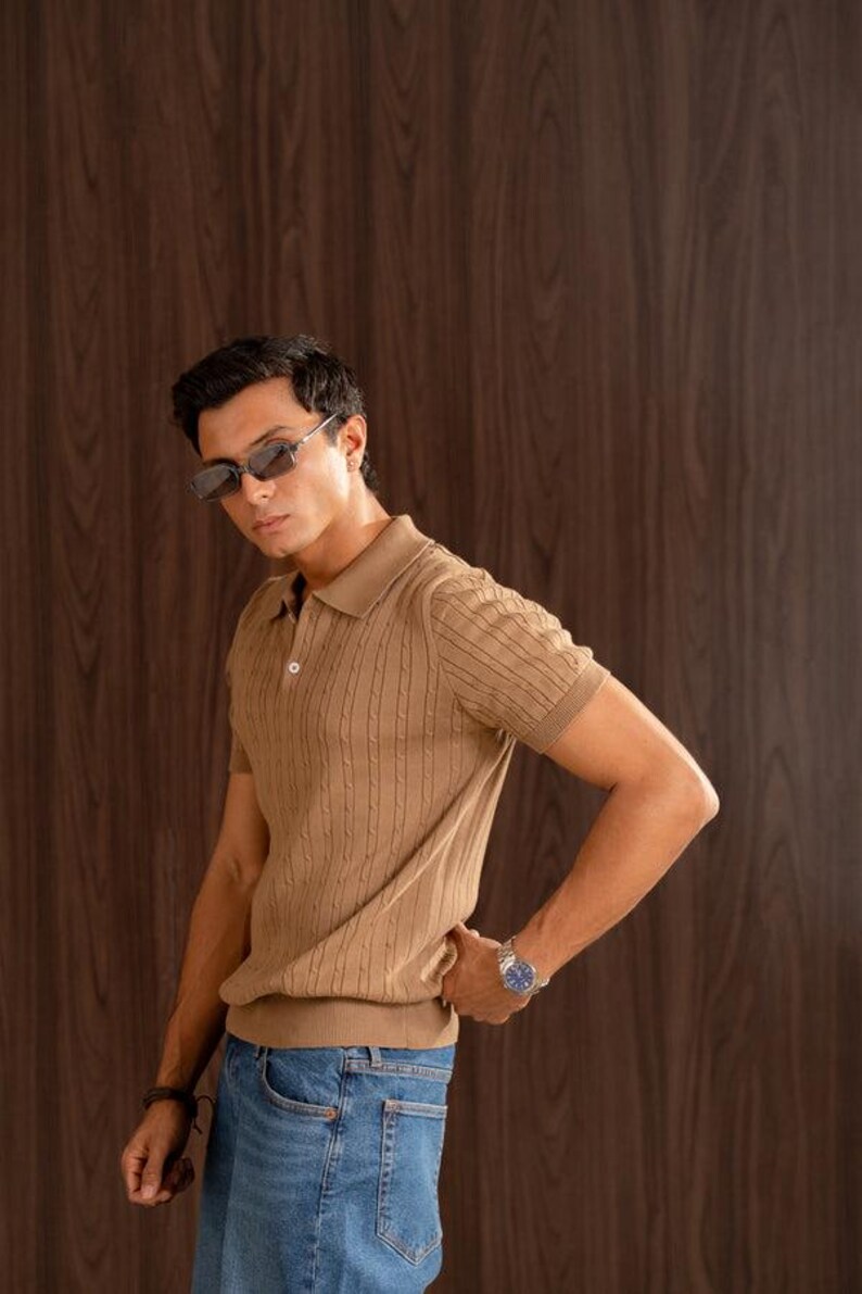 May include: A tan short-sleeved polo shirt with a textured cable knit design. The shirt has a collared neckline and a button closure. The model is wearing blue jeans and sunglasses. A watch is on the model's wrist.