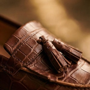 May include: A brown loafer shoe featuring a textured crocodile-embossed pattern across the upper surface. The shoe is detailed with a decorative leather strap across the vamp and two matching brown leather tassels attached to the front section.