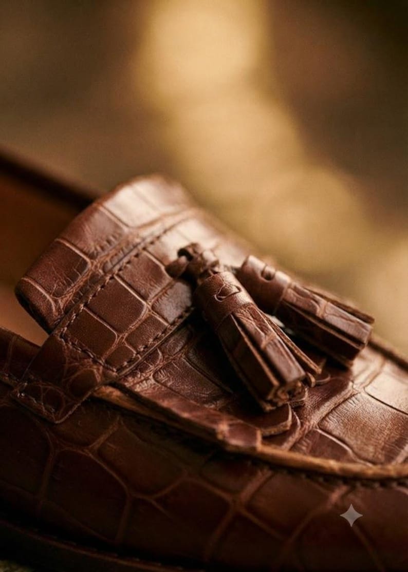 May include: A brown loafer shoe featuring a textured crocodile-embossed pattern across the upper surface. The shoe is detailed with a decorative leather strap across the vamp and two matching brown leather tassels attached to the front section.