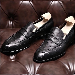 Pu&ograve; includere: Un paio di mocassini neri in pelle con superficie strutturata. Le scarpe slip-on sono presentate su una superficie in pelle marrone, con forme di legno all'interno.
