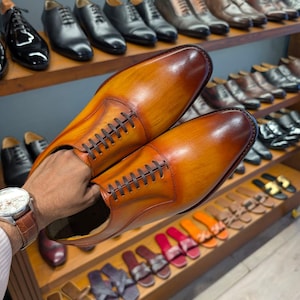 May include: A pair of brown leather dress shoes with a gradient finish, held in front of a display of various men's footwear. The shoes have a classic lace-up design and are displayed with other dress shoes and sandals.