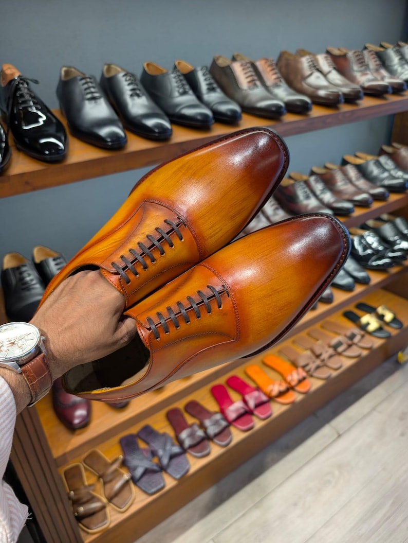 May include: A pair of brown leather dress shoes with a gradient finish, held in front of a display of various men's footwear. The shoes have a classic lace-up design and are displayed with other dress shoes and sandals.
