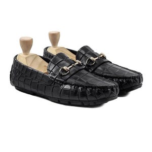 May include: A pair of black leather loafers with a crocodile texture. The shoes have a gold-colored metal accent across the top and are displayed with wooden shoe trees. The loafers are designed for a classic, sophisticated look.